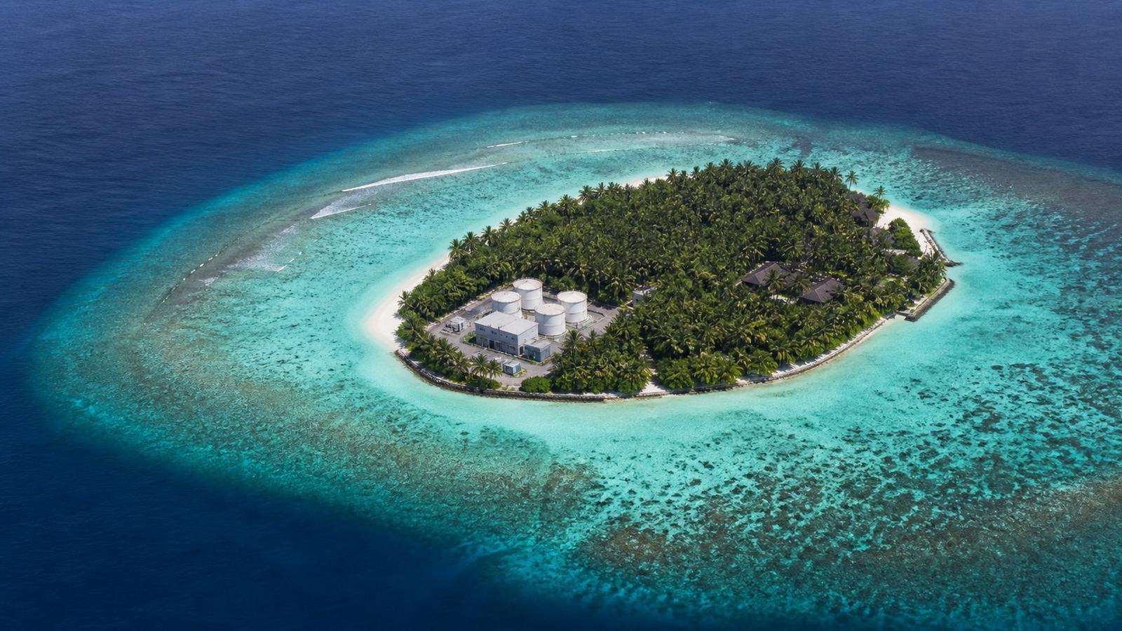Aerial view of a small Maldivian island ringed by a turquoise lagoon with water storage tanks visible